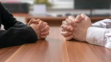 Deux personnes se font face à une table, mains croisées.